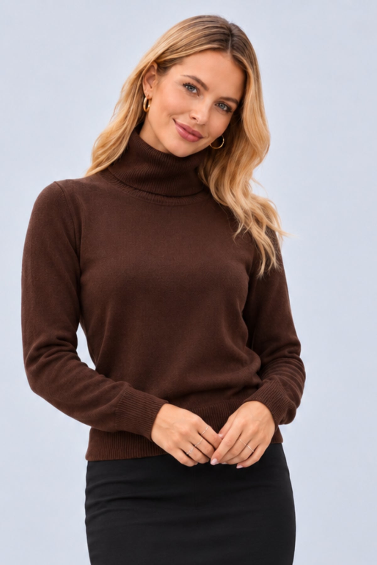 A woman wearing a brown ribbed turtleneck sweater and a black skirt, posing against a light gray studio background.