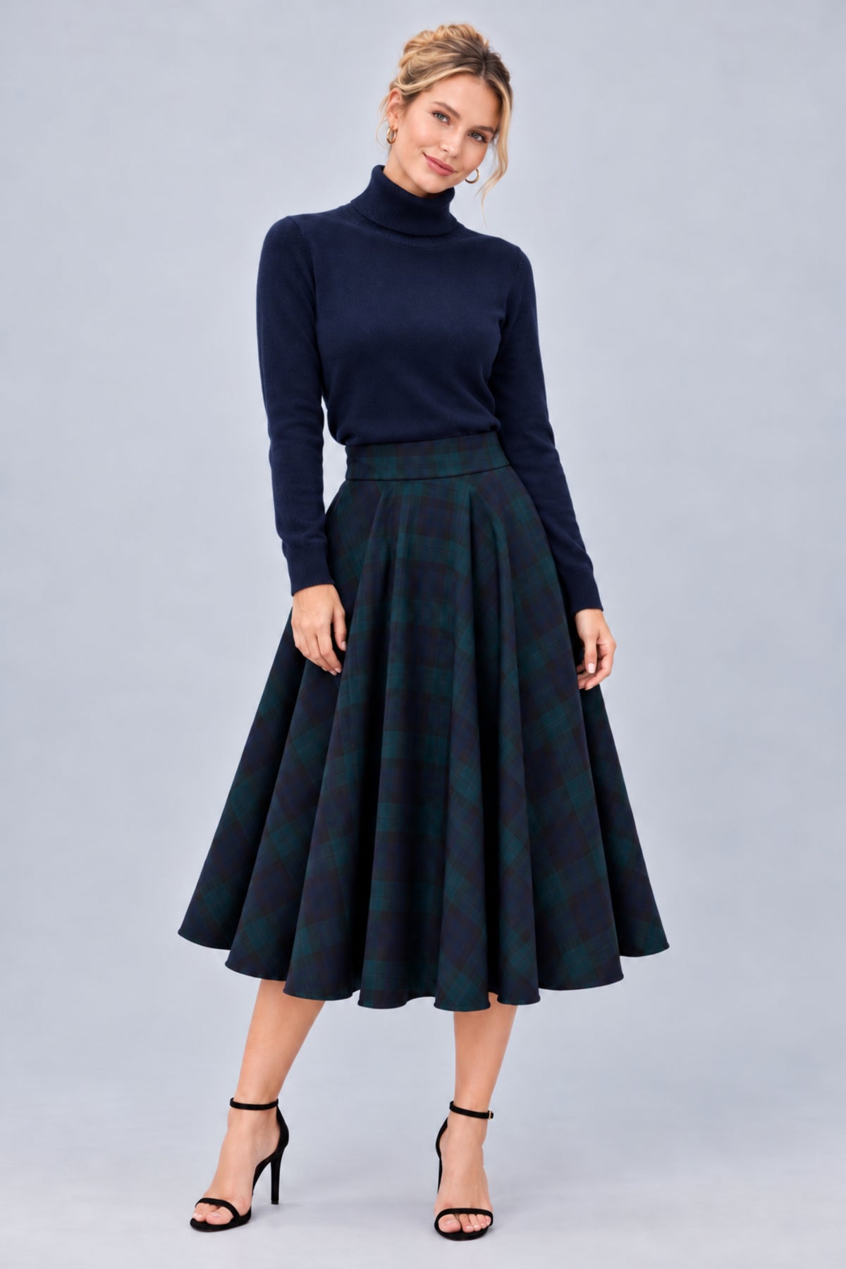 A woman modeling a dark navy blue mock neck knit top and a high-waisted, flared A-line skirt in a dark green and navy tartan plaid pattern, paired with black strappy high heels, against a solid light gray studio background.