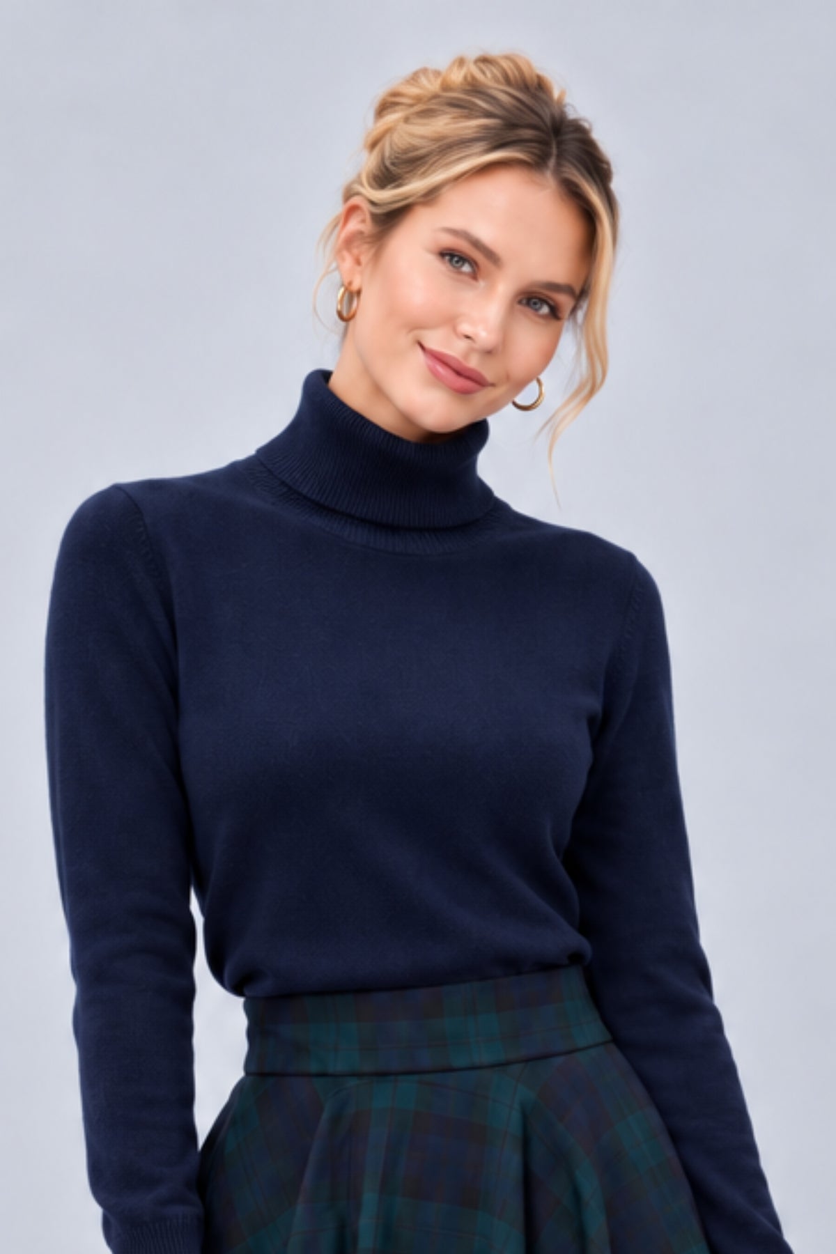 A woman wearing a dark navy blue turtleneck sweater and a green plaid skirt, posing against a solid light gray studio background.